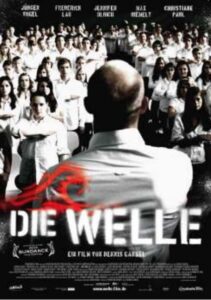 Bild zeigt das Filmplakat des Filmes die Welle. Wir sehen den Rücken des Lehrers. Er steht vor einer großen SchülerInnen-Gruppe in weißen Hemden. Sie alle zeigen den Gruß der Welle.